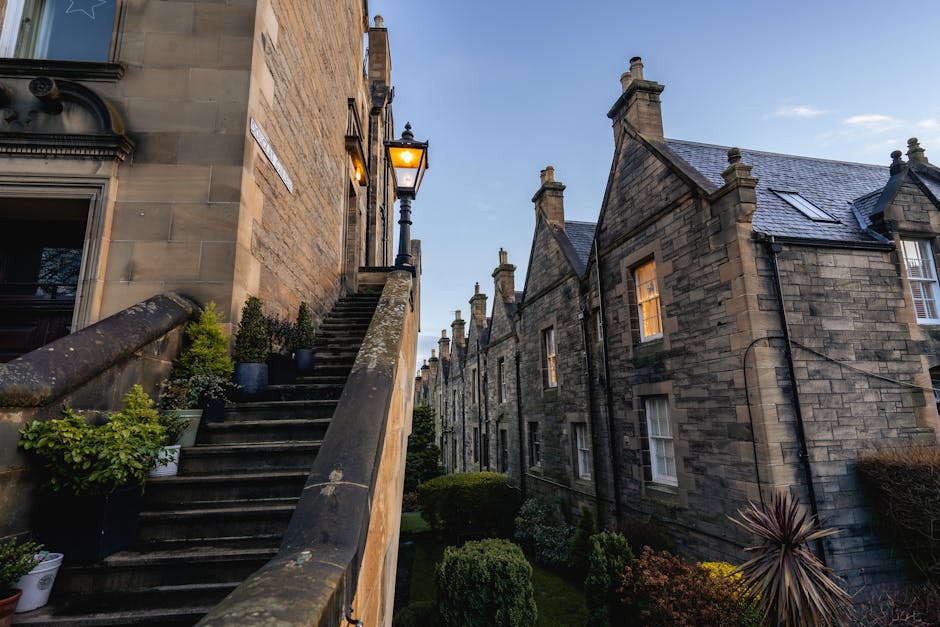 A wide city street in Edinburgh lined with multi-storey stone buildings, some under scaffolding with protective netting. Parked cars, including a black sedan and a white hatchback, are positioned alon