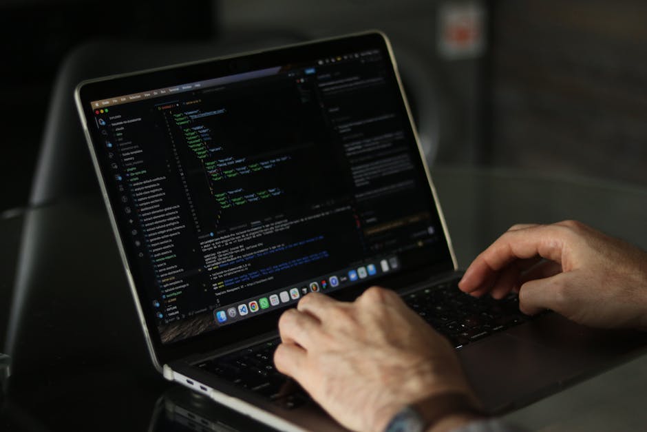 Close-up of a person's hands operating a laptop computer on a dark surface, with the screen displaying lines of code and text editor windows. The background is blurred, emphasizing the focus on the ha