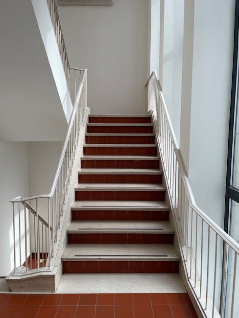 A straight staircase inside a residential building with white metal railings and wooden handrails on both sides, leading upward to an upper floor. The staircase has brown tiled treads and white risers